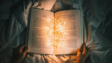 Open book held in hands with warm fairy lights resting on its pages on a cozy bed.