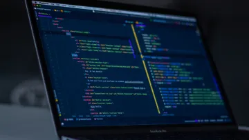 Laptop screen displaying colorful HTML and JavaScript code in a dark-themed development environment.