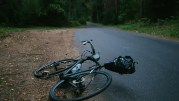 Black bicycle with a saddlebag lying on a forest road beside trees and soil under dim light.