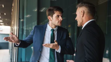 Two businessmen in suits engaged in a serious discussion with expressive gestures in an office corridor.