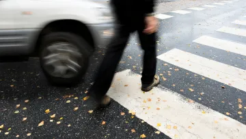 Blurred pedestrian walking across wet crosswalk while a silver car passes nearby on rainy street