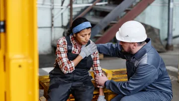 Male worker in white helmet comforting female worker sitting on pallet in industrial workplace with discomfort.