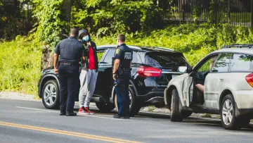 Two police officers talk to a man wearing a face mask beside parked cars on a roadside with greenery.