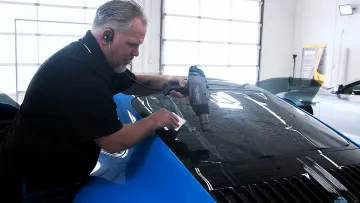 Man using heat gun to apply tinted film on blue car window in bright garage