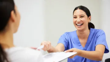Smiling healthcare professional in blue scrubs handing a clipboard to a patient in a medical setting.