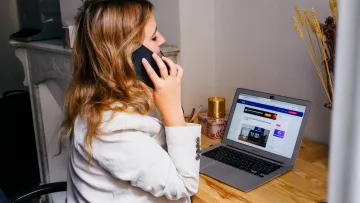 Woman in white blazer using smartphone while working on laptop at wooden desk in home office.