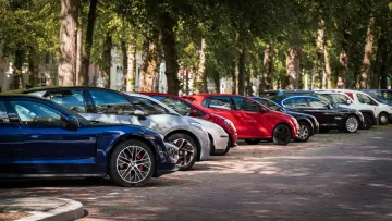 Various parked cars including blue, silver, red, and black models along a tree-lined street in daylight.