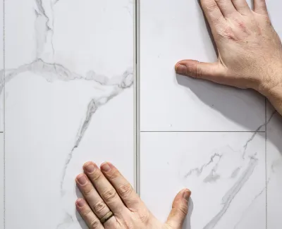 Two hands installing white marble-patterned ceramic tiles on a wall with gray grout lines