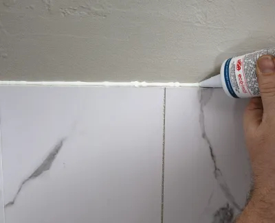 Applying white caulk along the seam between a gray wall and marble-patterned tile for finishing.