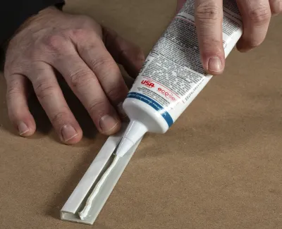 Hands applying white adhesive sealant from a tube onto a corner of a plastic or metal piece on a brown surface.