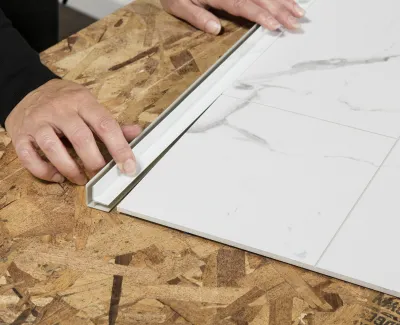 Hands aligning white marble-patterned tile with tile trim on plywood subfloor during installation.