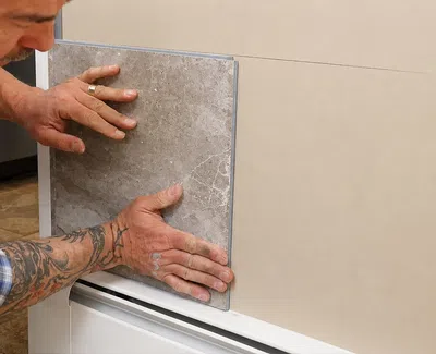 Man with tattoos installing large gray ceramic tile on indoor wall close-up.