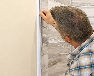 Man installing trim or molding on wall corner in a room with tiled wall and window, wearing plaid shirt.