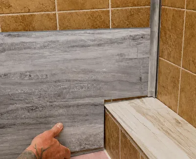 Hands installing a grey tile with a notch on a wall corner above a beige tiled step in a bathroom.