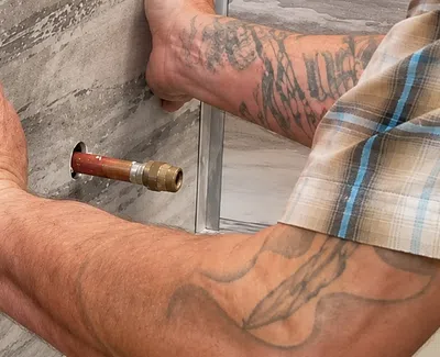 Man with tattoos wearing plaid shirt presses hand on wall near exposed copper pipe in construction setting