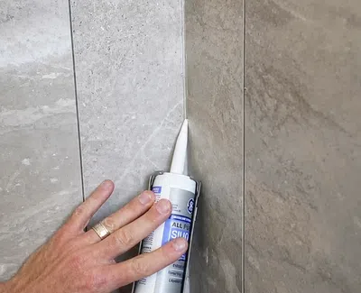 Hand applying white silicone sealant along the corner joint of tiled bathroom wall.