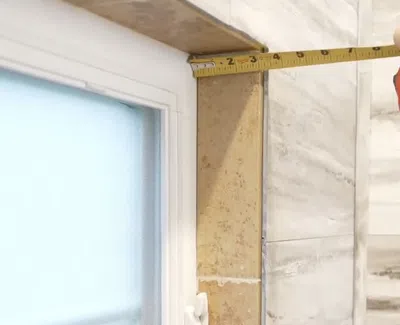 Hand measuring the width of a tiled wall corner near a window using a tape measure.