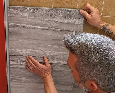 Man installing large gray tile on a wall next to a red surface with a plumbing fixture opening.