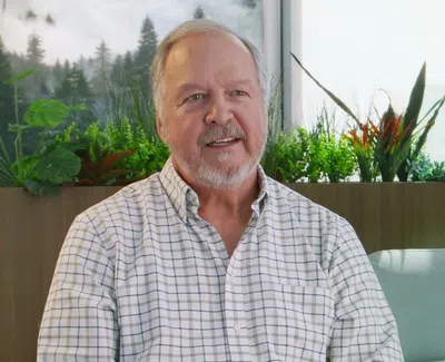 Older man with beard wearing a checkered shirt sitting on a couch with indoor plants in the background