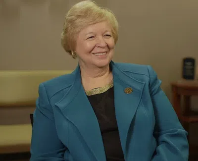 Smiling older woman in blue blazer sitting in a warmly lit room with chairs and a small table in the background