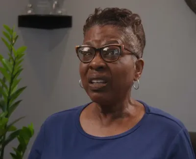 Older woman with glasses and hoop earrings looks concerned while sitting indoors next to a green plant.