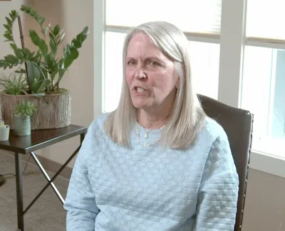 Middle-aged woman with long gray hair sitting in a chair in a bright room with plants and a sofa.