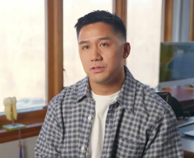 Man in a checkered shirt sits in a bright room with large windows, plants, and a computer in the background.