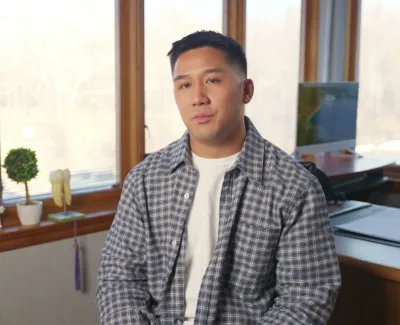 Man in checkered shirt sitting in bright office with large windows and desk in background