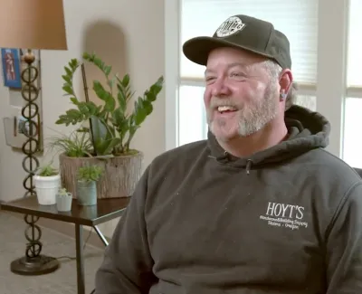 Smiling man in casual hoodie and cap sitting indoors near plants and window with natural light.