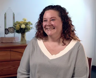 Smiling woman with curly hair sitting in front of wooden cabinet with plants and flowers in background
