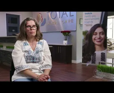 Woman with glasses sitting in a dental office with Total Dental Care sign and Invisalign poster in background