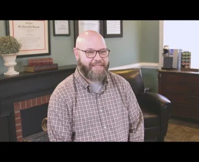 Smiling bearded man with glasses in plaid shirt sitting in cozy office with certificates and bookshelf.