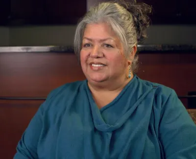 Smiling woman with gray hair wearing teal sweater seated indoors against a blurred background.