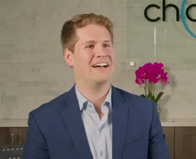 Smiling young man in blue blazer sitting in modern dental office with pink orchids and logo on wall.