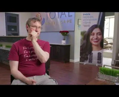 Man in maroon shirt sitting and covering mouth with hand in a modern office with promotional posters.