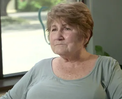 Elderly woman with short hair sitting indoors near a window, looking thoughtfully to the side.