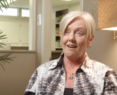 Smiling senior woman with short blonde hair wearing a patterned shirt and cross necklace in a cozy home setting