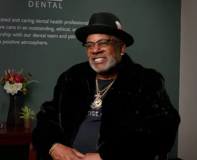 Smiling man in black hat and fur coat sitting in a dental office with flowers and award on table