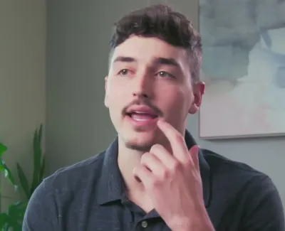 Man with short curly hair and mustache speaking indoors near a window with green plants and abstract wall art.