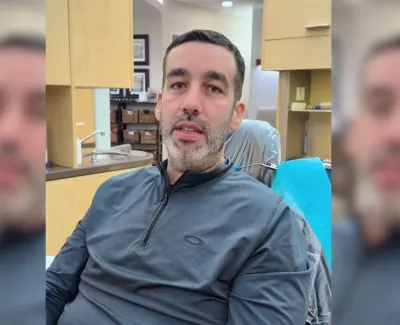 Man with short dark hair and beard wearing gray zip-up jacket sitting indoors at a desk area.