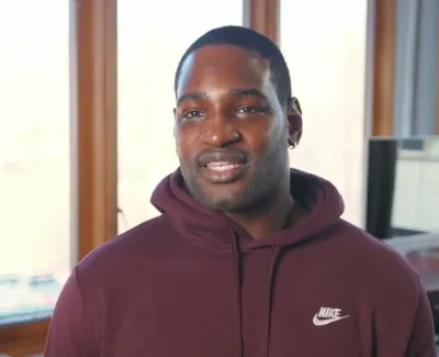 Smiling man wearing a maroon Nike hoodie in a well-lit room with wooden window frames and a small plant on the windowsill.