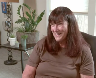 Smiling woman with brown hair seated in a cozy living room with plants and natural light.