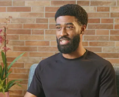 Man with beard sitting indoors near plants against a brick wall, smiling and wearing a black t-shirt.