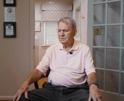 Elderly man sitting in a chair in a well-lit room with window blinds and framed signs on the wall.
