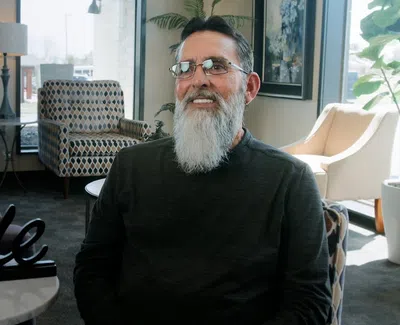 Smiling elderly man with glasses and white beard sitting in a cozy, well-lit living room with modern furniture.