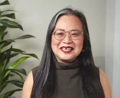 Smiling woman with glasses and long hair sitting near a large green plant and a motivational sign
