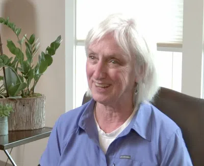 Smiling older woman with white hair sitting indoors near window and green potted plants on side table.