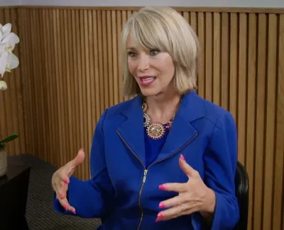 Woman in blue jacket talking and gesturing in an indoor setting with wooden panel and white orchid plant.