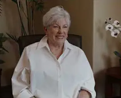 Smiling elderly woman with white hair sitting in a chair in a warmly lit room with plants and flowers.