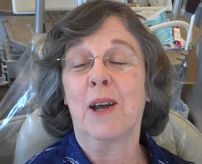 Elderly woman with glasses sitting in a dental chair with eyes closed and mouth slightly open.